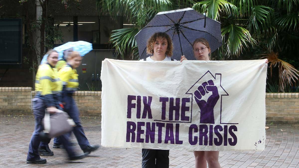  Two women hold a banner that says "FIX THE RENTAL CRISIS" in front of a brick building.